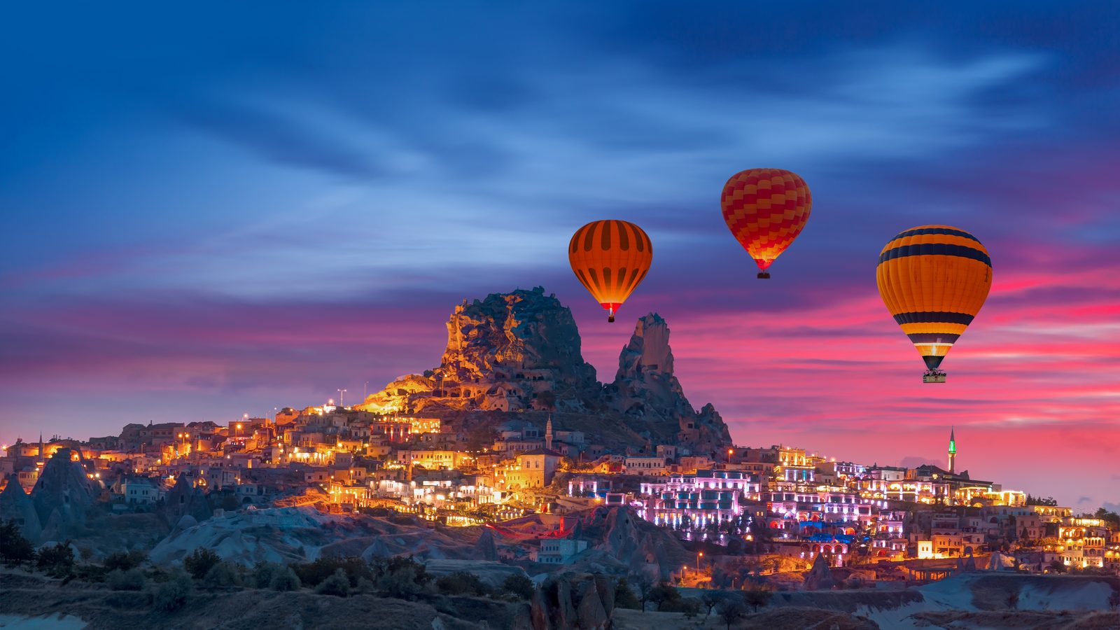 Hot-air balloons rising over Cappadocia at dusk, with the lit town below