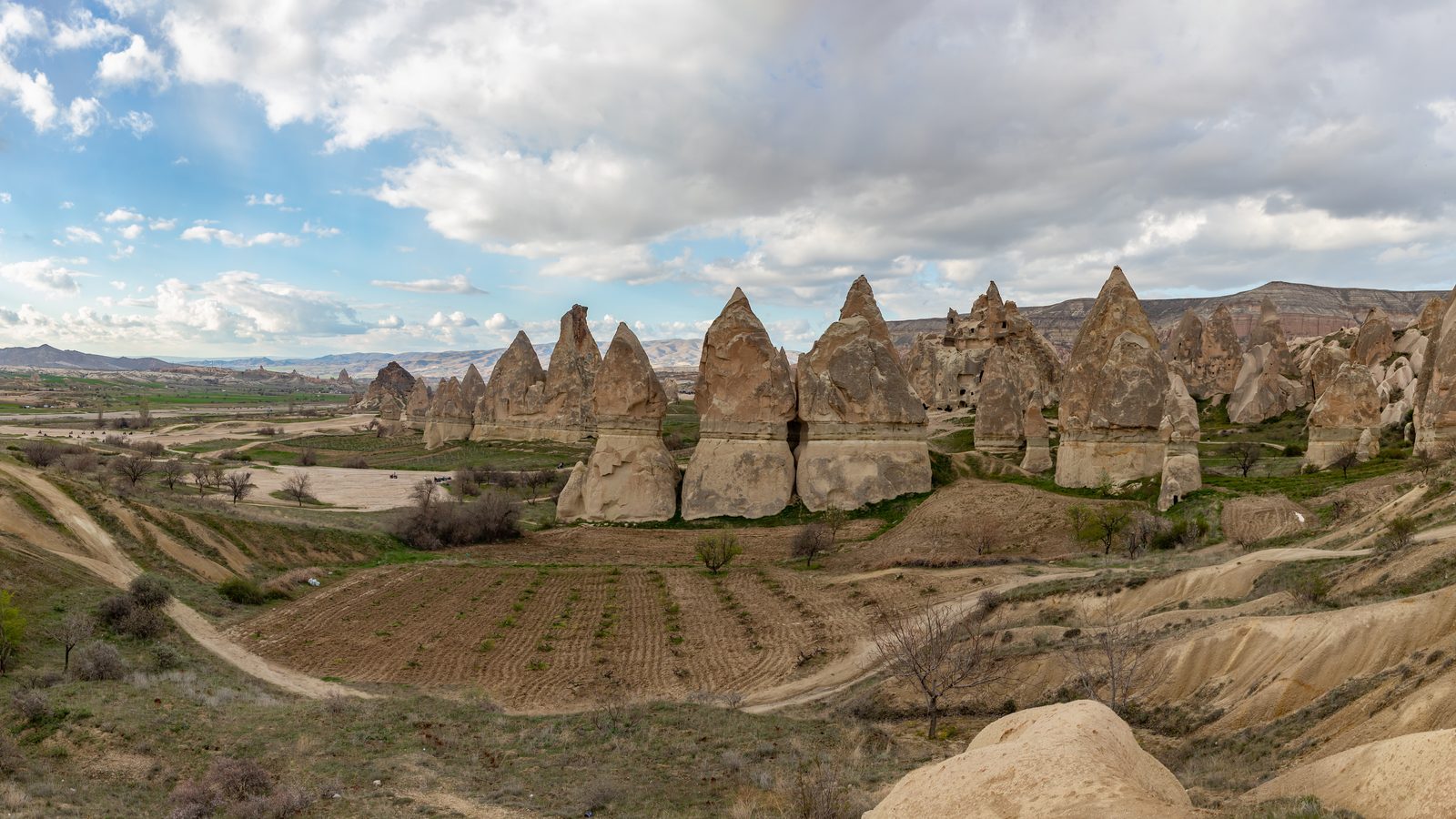 Cone-shaped fairy chimneys above a ploughed valley floor near Göreme, under a broken spring sky