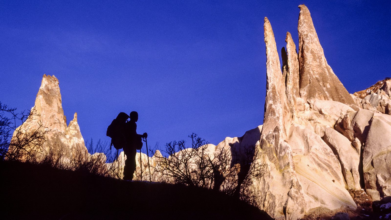 A walker with poles silhouetted against sunlit fairy chimneys under a deep blue sky, patches of snow on the ground
